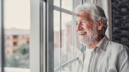 Eine Person mit kurzen, weißen Haaren und weißem Bart steht direkt neben einer Terrassentür. Sie spiegelt sich im Glas. Die Person lächelt