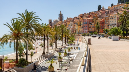 Eine Küstenstadt mit dicht gebauten, farbigen Häusern an einem Hang. Im Vordergrund ist eine breite Promenade mit Geländer, daneben ein Strand mit einzelnen Personen. Das Meer liegt links im Bild, während rechts die Promenade weiterführt