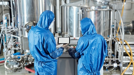 Zwei Personen in blauen Schutzanzügen in Rückenansicht stehen vor Steuerungsanlage, im Hintergrund Metallfässer und Leitungen