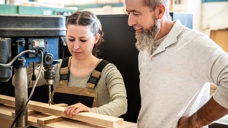 Zwei Personen unterschiedlichen Alters stehen in einer Tischlerei bei einer Bohrmaschine. Die jüngere Person blickt konzentriert auf ein Werkstück auf dem der Bohrer angesetzt ist. Die ältere Person schaut dabei zu. Beide wirken zufrieden.