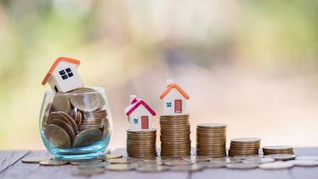 Auf einer hölzernen Oberfläche stehen direkt nebeneinander fünf unterschiedlich hohe Münzstapel. Links daneben ist ein kleines, durchsichtiges Glas, in dem weitere Münzen sind, darauf und auf zwei Stapeln stehen kleine Miniaturhäuser.