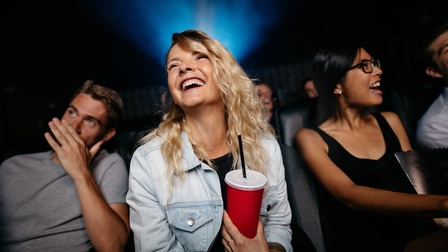 Vier Personen sitzen auf Sesseln mit hohen Rückenlehnen nebeneinander. Eine Person hält einen roten Becher mit schwarzem Strohhalm und lacht. Dahinter sitzen weitere Personen in einer Reihe.