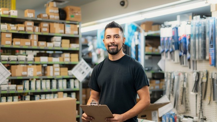 Halbporträt einer Person mit kurzen, dunklen Haaren und Bart, die ein Klemmbrett hält. Im Hintergrund hängen verpackte Werkzeuge an einer Wand sowie ein mit Kartons befülltes Regal.