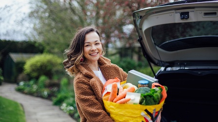Frontalaufnahme einer Person, die mit beiden Händen eine große, gelbe Tüte hält, in der unter anderem Karotten, Brokkoli oder eine Tetrapaktüte sind. Neben der Person parkt ein Auto mit einem geöffneten Kofferraum