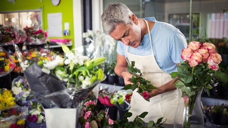 Frontale Aufnahme einer Person mit kurzen, grauen Haaren und Schürze, die an einem Tisch steht und eine Rose hält, auf die sie blickt. Um die Person viele weitere Blumen sowie verschiedene Blumensträuße
