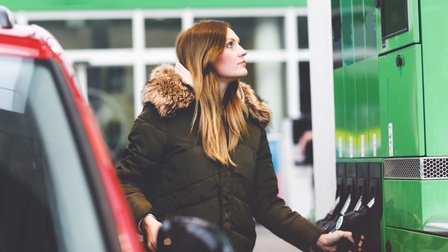 Eine Person in einer dicken Jacke und mit offenen Haaren steht an einer Zapfsäule und hält mit einer Hand einen Zapfhahn. Links neben ihr steht ein PKW