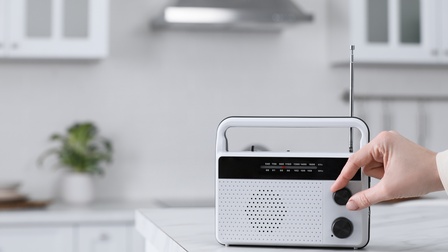 Ein Radio steht auf einem Tresen in einer hellen Küche. Die rechte Hand einer Person dreht an einem Rädchen des Radios