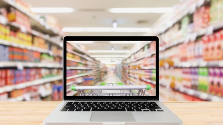 Frontalansicht eines aufgeklappten Laptops auf einer hölzernen Unterlage. Auf dem Bildschirm ist ein Einkaufswagen, der links und rechts von Regalen, befüllt mit Gegenständen, umgeben ist