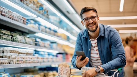 Eine Person mit Brille und Jeanshemd hinter einem Einkaufswagen im Supermarkt neben dem Kühlregal mit Milchprodukten und Eiern. Die Person hält ein Smartphone in der Hand und lächelt freudig. Im Einkaufswagen befinden sich einige Produkte.