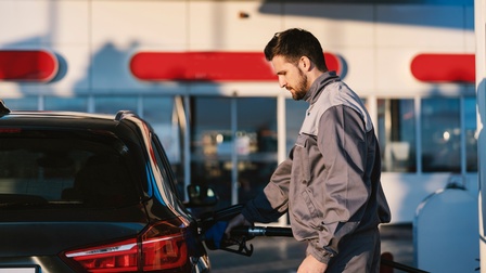 Eine Person in Arbeitskleidung steht an einem Auto auf einer Tankstelle und tankt das Gefährt.