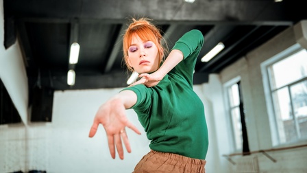 Eine Person mit orangen Haaren und lila Lidschatten steht in einem hohen Raum und tanzt. Der rechte Arm ist ausgestreckt. Der linke Arm ist angewinkelt.