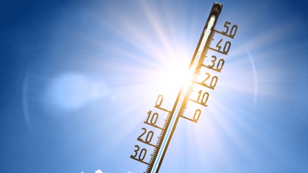Nahaufnahme eines Thermometers vor dem Hintergrund eines blauen Himmels mit ein paar Wolken. Rechts sind die Zahlen bis 50 daran montiert. Das Thermometer wird von strahlendem Sonnenlicht beleuchtet