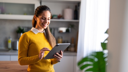 Nahaufnahme einer Person, die ein Tablet in der Hand hat, in das sie hineintippt. Im Hintergrund sieht man eine Küche mit Küchengeräten, außerdem sieht man eine Zimmerpflanze. Die Person lehnt mit der Hüfte auf einem Küchenblock aus dunkelbraunem Holz