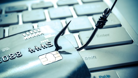 Auf einer Computertastatur liegt eine Kreditkarte, in der ein kleines Loch ist, durch das ein Fischhaken eingefädelt ist