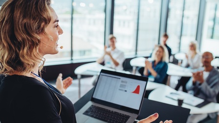 Seitlich Aufnahme einer Person mit Mikrofon vor dem Mund, die an einem Pult steht. Auf dem Pult ist ein aufgeklappter Laptop. Die Person blickt zu sechs Personen, die vor ihr an Tischen sitzen. Die Personen klatschen in die Hände