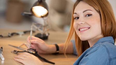 Lächelnde Person im Fokus, im Hintergrund verschwommen Tisch, an dem Person Uhr mit Werkzeugen unter Lampe bearbeitet
