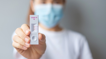 Person mit Mundnasenschutz hält Covid-Antigen-Test mit positivem Testergebnis