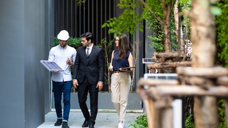 Drei Personen spazieren durch Grünanlage eines Gebäudes, eine Person trägt weißen Schutzhelm und hält kleines Solarpaneel in Hand, die anderen Personen blicken darauf