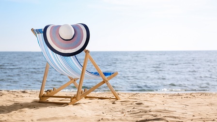 Rückenansicht eines Liegestuhls, der auf einem Sandstrand steht. Auf dem Liegestuhl ist ein Sonnenhut. Am Ende des Strands ist das Meer