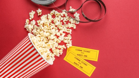 Popcornbehälter liegt auf einem roten Untergrund, daneben liegen zwei gelbe Kinotickets sowie eine Filmspule