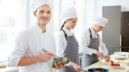 Drei Personen stehen bei einer Kücheninsel in weißer Arbeitskleidung mit Kochhauben und verarbeiten Lebensmittel, zwei Personen tragen eine grau-gestreifte Schürze