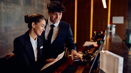 Seitliche Aufnahme zweier Personen in Anzügen, die hinter einem Tresen an einem Computer stehen. Die linke Person hat ein kleines Ausweisschild auf der Anzugjacke und hält einen Zettel. Die rechte Person deutet auf den Bildschirm