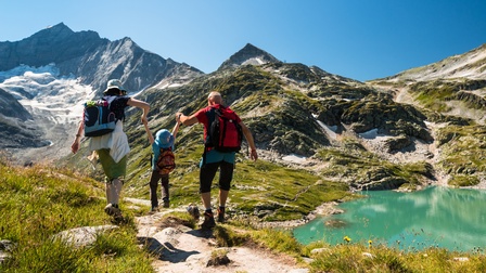 Zwei erwachsene Personen heben beim Wandern an beiden Händen Kind in die Luft, ringsum Berglandschaft, Wiesen und Wasser
