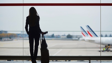 Person in Rückenansicht im Gegenlicht mit Tasche und Rollkoffer sowie Flugticket und Reisepass in Hand steht vor großem Fenster und blickt auf Rollbahn mit Flugzeugen