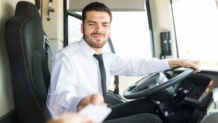 Person mit kurzen dunklen Haaren, Bart sowie weißem Hemd und Krawatte sitzt am Steuer eines Fahrzeuges und nimmt eine Karte entgegen