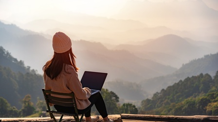 Person mit Mütze in Rückenansicht sitzt auf Sessel mit aufgeklapptem Notebook am Schoß und blickt auf Berg- und Hügelpanorama vor sich