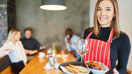 Lächelnde Person mit rot-weiß gestreifter Schürze hält Tablet mit gefülltem Weißweinglas und zwei Tellern mit Speisen in Händen, im Hintergrund verschwommen drei Personen an Tisch sitzend