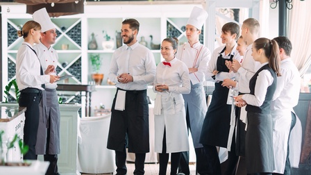 Mehrere Personen in Gastronomiegewändern, wie Schürzen und Kochhauben, stehen in Restaurant und blicken auf zwei weitere Personen ihnen gegenüber stehend
