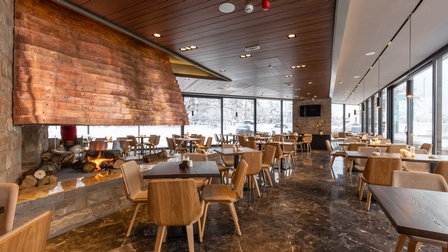 Blick auf einen Restaurantbereich mit offener Feuerstelle und vollflächigen Fensterfronten mit Blick auf eine Winterlandschaft