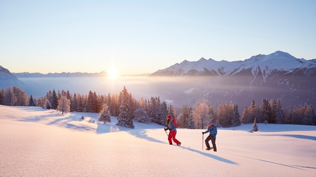 Zwei Personen im Sonnenuntergang in Wintergewändern mit Stecken und Schneeschuhen wandern durch verschneite Landschaft, im Hintergrund beschneite Bäume und Berge