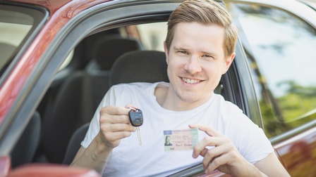 Lächelnde Person lehnt sich auf Fahrerseite aus Auto und hält Führerschein und Autoschlüssel in Händen