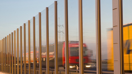 Transparente Lärmschutzwand in Sonnenlicht, durch die LKWs auf Autobahn verschwommen erkennbar sind