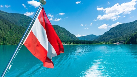 Österreichflagge weht im Wind während ein Boot entlang eines Seengebietes in malerischer Bergkulisse entlangfährt