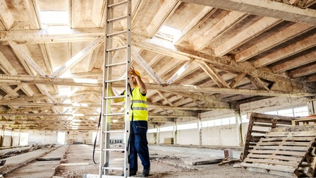 Eine Person mit Helm und gelber Schutzweste steht in einer großen leeren Halle neben einer sehr hohen Leiter und hält diese mit den Händen fest. Rechts neben der Person liegen Paletten. Am Boden liegt Schutt