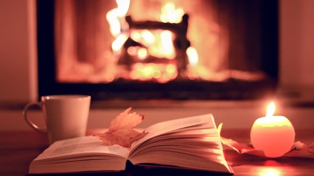 Aufgeklapptes Buch mit gelb gefärbtem herbstlichen Blatt darauf liegend, neben brennender runder Kerze und Tasse, im Hintergrund Kaminfeuer
