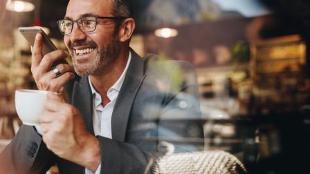 Lächelnde Person in Anzug mit breitem Silberring hält Smartphone zum Mund und in anderer Hand Kaffeetasse
