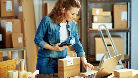 Eine Person steht in einem Warenlager und hält ein Handy in der Hand. Sie blickt auf ihren Laptop und tippt. Auf dem Tisch befinden sich Kartons und Klebematerial. Im Hintergrund sind Regale mit Kartons zu erkennen.
