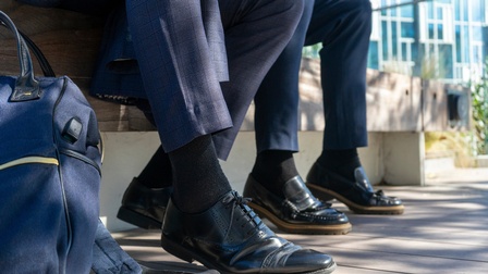Nahaufnahme von Füßen von Personen in Businesskleidung mit schwarzen Socken und Lackschuhen, im Hintergrund steht ein Gebäude mit Glasfronten