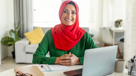 Porträt einer lächelnden Person mit rotem Kopftuch vor aufgeklapptem Laptop am Schreibtisch sitzend, ringsum Büroutensilien