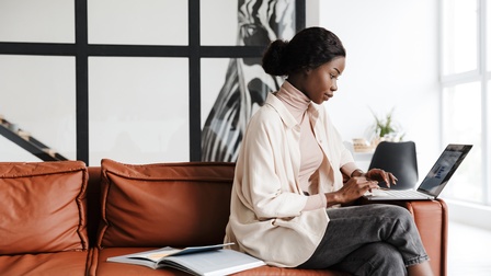 Person mit geschlossenen schwarzen Haaren und Pullover, Bluse und Jeans sitzt bei einer Couch und arbeitet mit einem Laptop