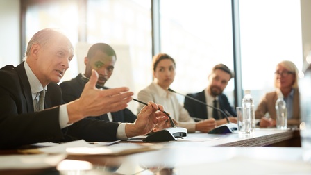 An einem Tisch sitzen mehrere Personen in Sakkos. Vor ihnen sind teilweise Mikrofone. Eine der Personen spricht in ein Mikrofon. Die anderen blicken zu der Person