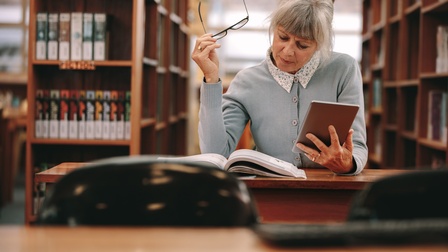 Person sitzt in Bibliothek und blickt auf Buch, hält in einer Hand eine Brille und in der anderen ein Tablet