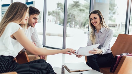 Um einen niedrigen Tisch sitzen drei Personen. Die Person rechts hält den beiden Personen links einen Zettel hin, auf den alle drei blicken. Im Hintergrund ist eine Glasfront