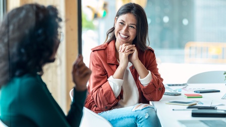 Lächelnde Person mit langen dunkeln Haaren hält beide ineinandergreifende Hände ans Kinn, im Vordergrund unscharf weitere Person