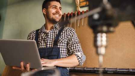 Lächelnde, bärtige Person in Arbeitslatzhose blickt zur Seite und hält aufgeklapptes Notebook in Händen, im Hintergrund Panel an Wand mit rotschwarzen Schraubenziehern unterschiedlicher Größen, im Vordergrund verschwommen Drechselbohrer