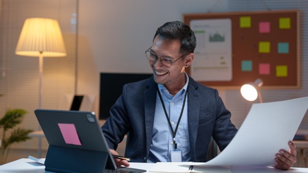 Eine Person mit Sakko, Ausweis um den Hals sowie Brille sitzt an einem Schreibtisch. In der linken Hand hält sie einen großen Zettel. Mit der rechten Hand bedient sie ein aufgestelltes Tablet